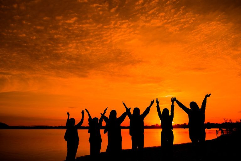 A group of friends enjoy the sunset by the lakeshore, creating beautiful silhouettes against the orange sky.