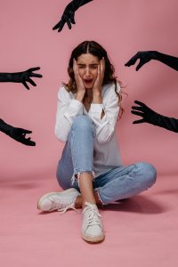 A young woman sits frightened as dark gloved hands reach towards her against a pink background.