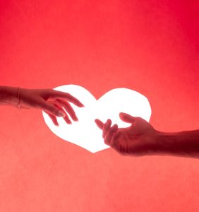 Two hands reaching towards each other over a heart shape on a vivid red background, symbolizing love and connection.