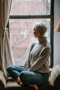 Serene woman in casual attire sitting on windowsill, gazing outside, enjoying peaceful solitude. 
