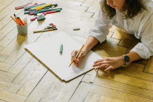 Woman lying on wooden floor drawing with pencil and paper surrounded by colorful stationery tools.