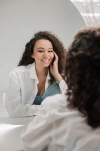 A woman in a white shirt smiles at her reflection in a mirror, exuding happiness and self-love.
